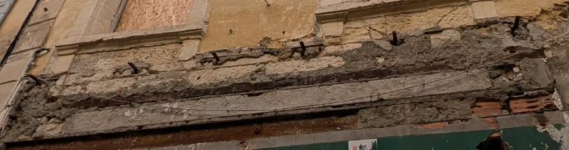 Bandeau de façade dégradé : enduit tombé, maçonnerie à nu et fers corrodés au-dessus d'un rez-de-chaussée.